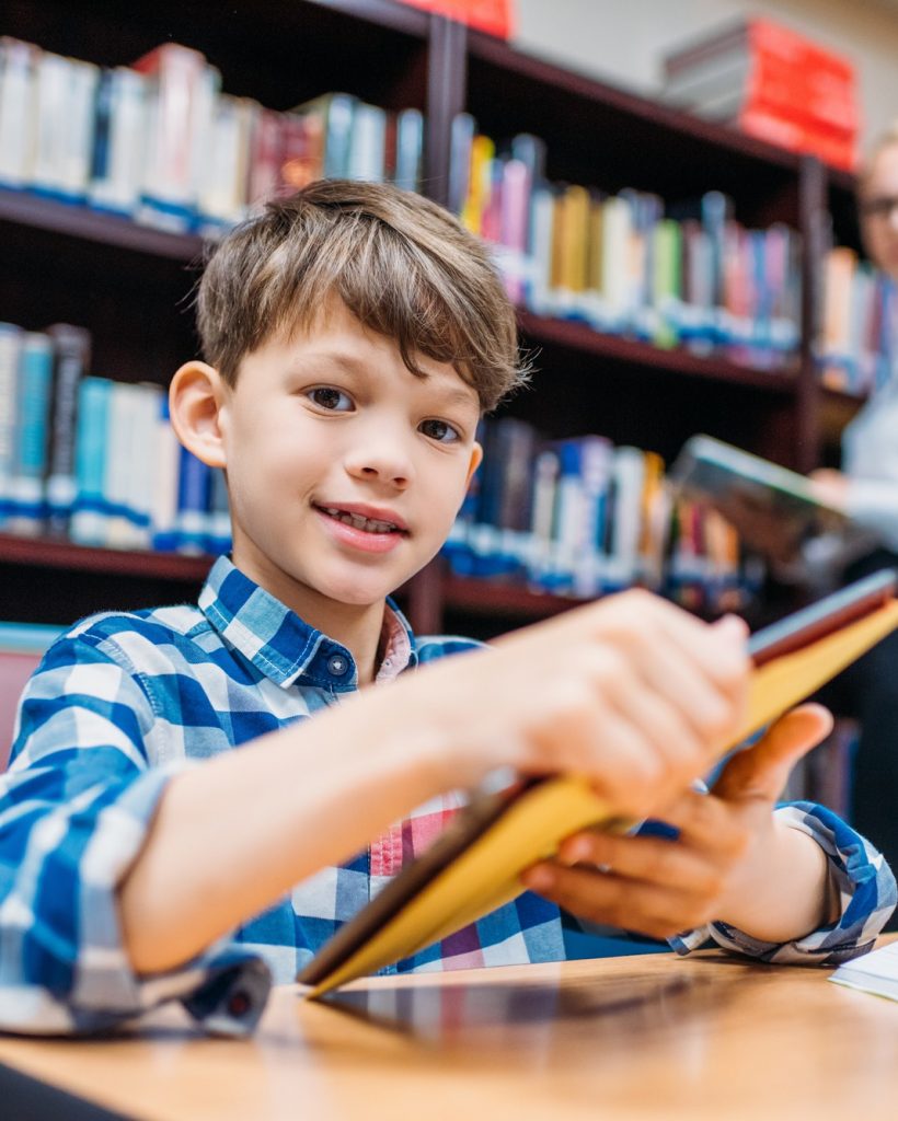 cute schoolboy holding tablet in library with teacher blurred on backgound